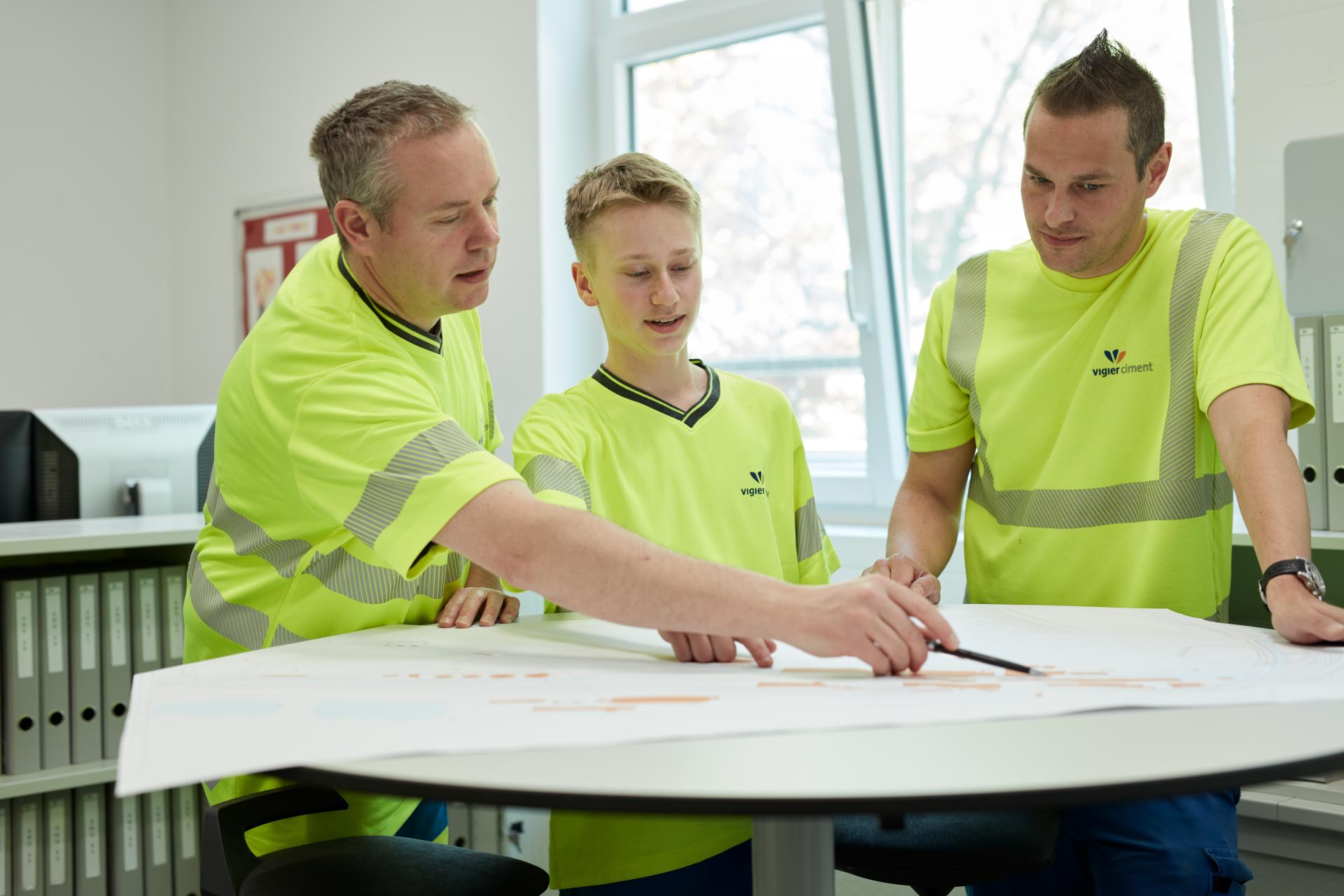 Mehrere Mitarbeiter diskutieren einen Plan mit einem Lehrling. Sie tragen Leuchtkleidung.